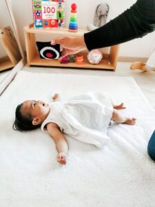 Un bebé con un vestido blanco yace sobre una colchoneta suave, mirando hacia un cojín sensorial para bebés. Cerca hay estanterías con juguetes y libros coloridos.
