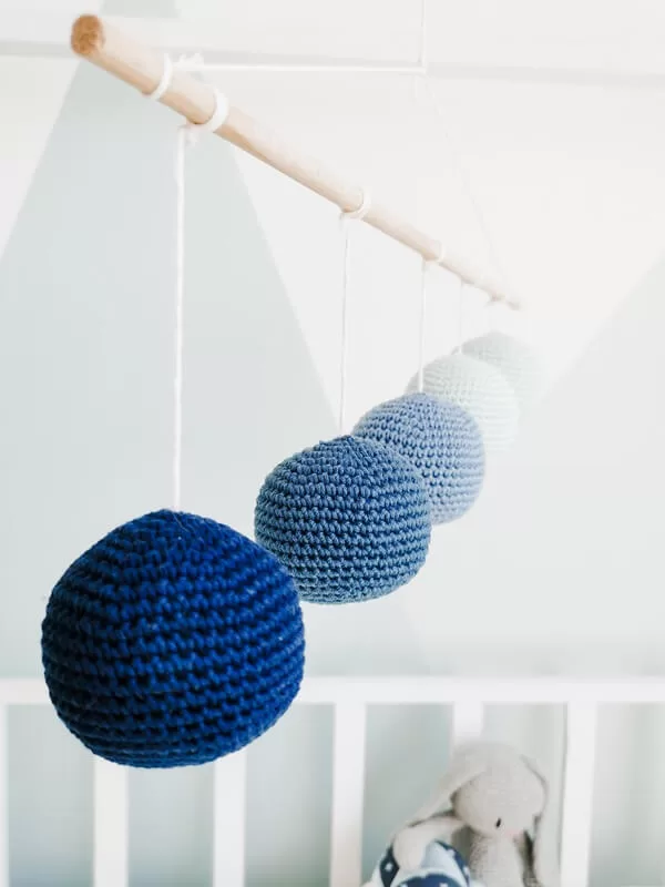 Móvil para bebé estilo Montessori con esferas de crochet en tonos degradados de azul, colgado de una varilla de madera. Se encuentra sobre una cuna blanca con un peluche de fondo.