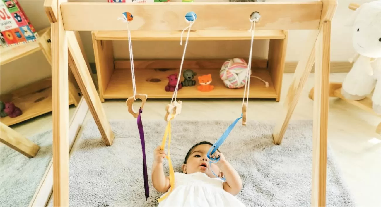 Bebé acostado boca arriba en un gimnasio de madera Montessori, sujetando una cinta azul con un mordedero de madera en forma de oso. Otros dos colgantes con cintas morada y amarilla cuelgan del gimnasio. En el fondo, juguetes, libros y una estantería de madera complementan el ambiente cálido y estimulante.