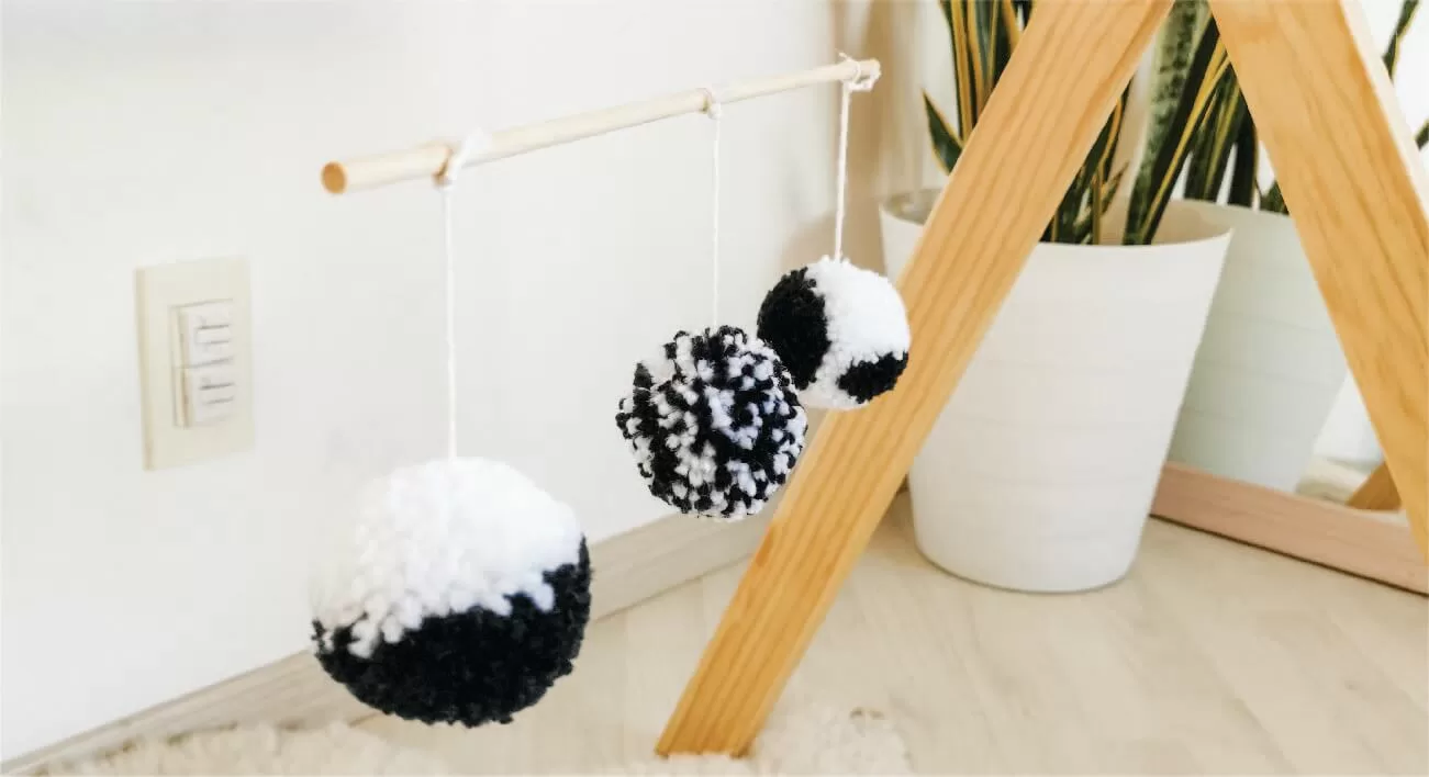 tres pompones en alto contraste blanco y negro de diferentes formas cuelgan de una bara de madera. Detras se observa una planta