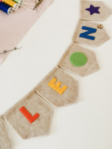 Banderines de fieltro con el nombre "LEON" en letras de colores vibrantes. Incluye una estrella morada, un botón decorativo y un círculo verde como detalles. En el fondo se observan carretes de hilo y agujas, destacando el proceso artesanal de su elaboración.