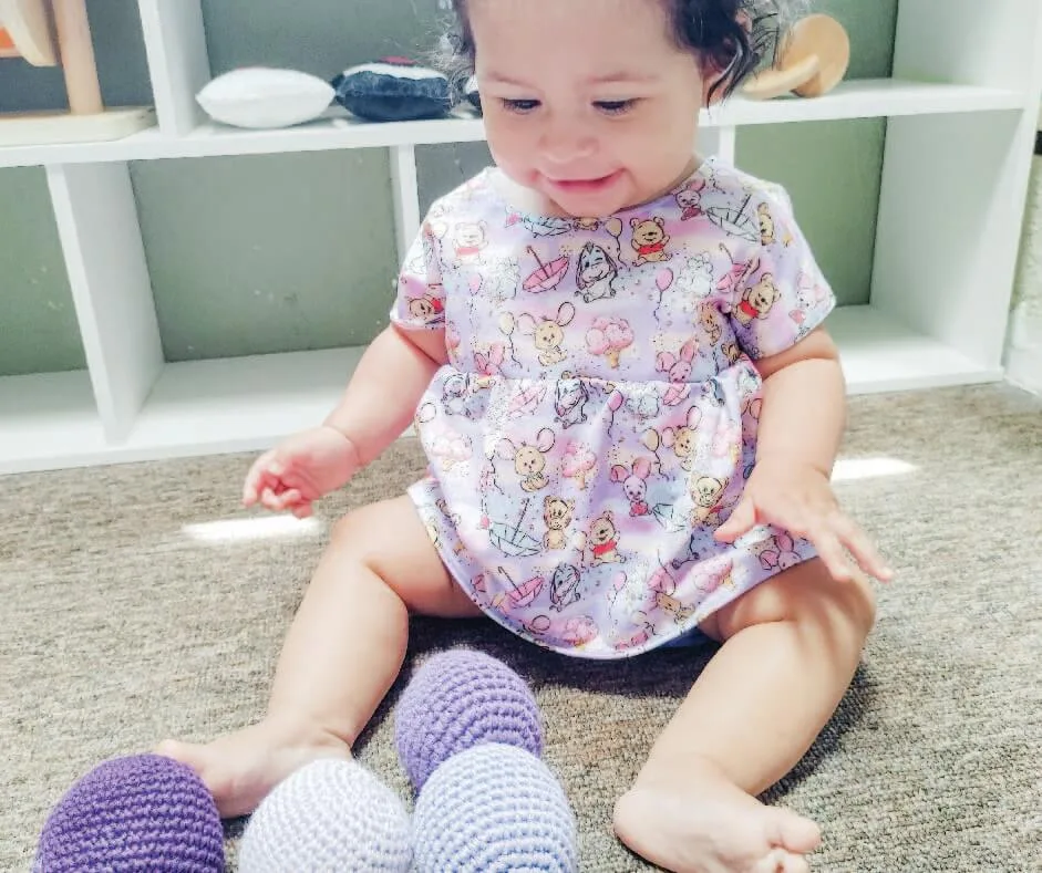 Bebé sentado en el suelo, mirando esferas de crochet color morado. En el fondo se aprecia un shelf montessori de color blanco