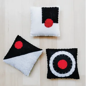 Tres cojines sensoriales Montessori de fieltro en colores blanco, negro y rojo, con costuras visibles y patrones geométricos contrastantes, colocados sobre una superficie de madera clara.