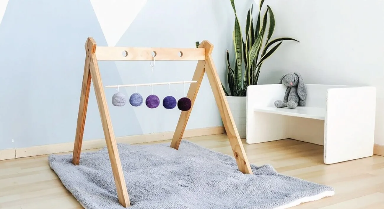 Móvil Gobbi Montessori colgado en un gimnasio de madera natural. Las esferas de crochet en tonos degradados de morado cuelgan de una barra delgada, diseñadas para estimular el desarrollo visual del bebé. La escena se sitúa en una habitación infantil con paredes en tonos pastel, una alfombra mullida y un sillón blanco con un peluche gris en el fondo.