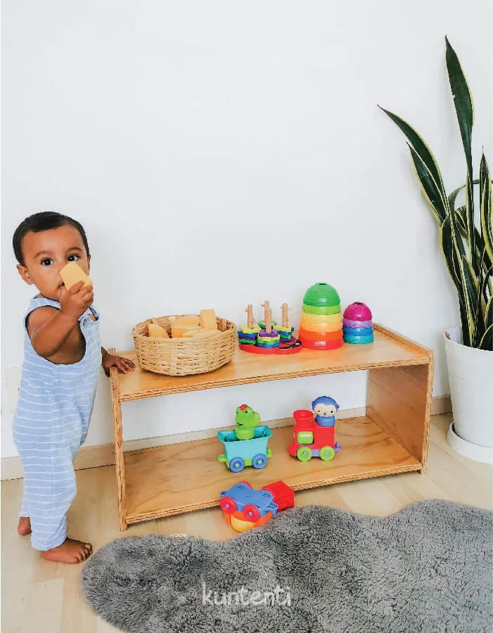Bebe sostieniendose de shelf montessori mientras toma una pieza de madera y la enseña. En el mueble montessori se ven distitnos materiales para bebes de colores vibrantes. Junto al shelf se ve una planta.