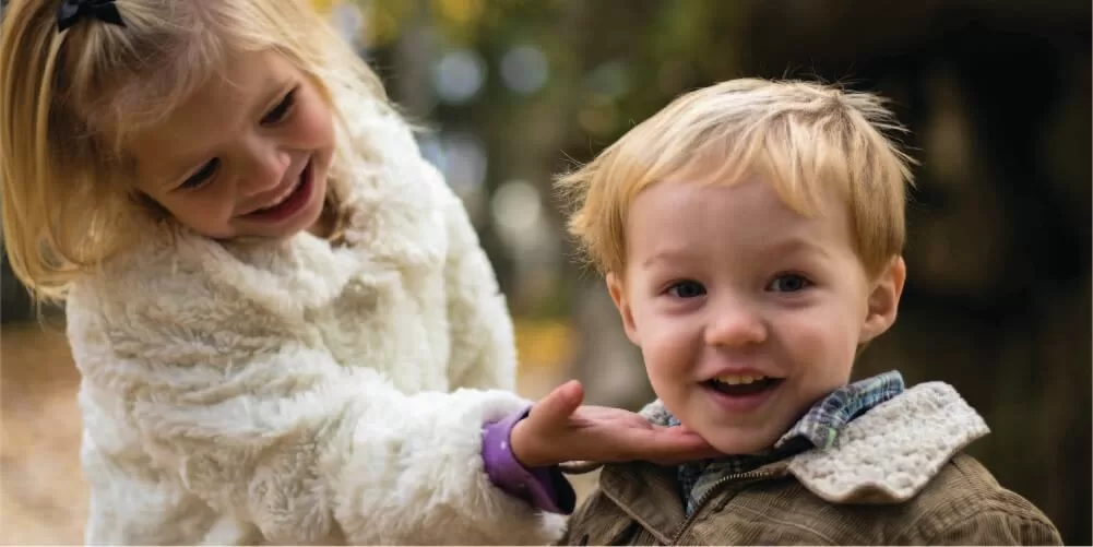 una niña pequeña acaricia la barbilla de un niño pequeño que sonríe, detrás de ellos se ve un bosque