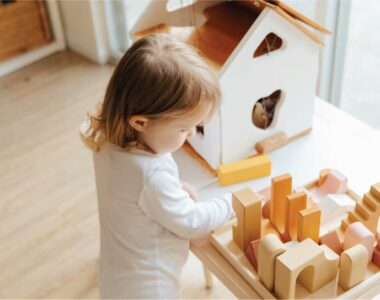 Niña pequeña jugando con bloques de construcción de madera en una mesa Montessori, mientras explora libremente en un ambiente preparado. A su lado, una casita de madera refuerza el juego simbólico y la autonomía.