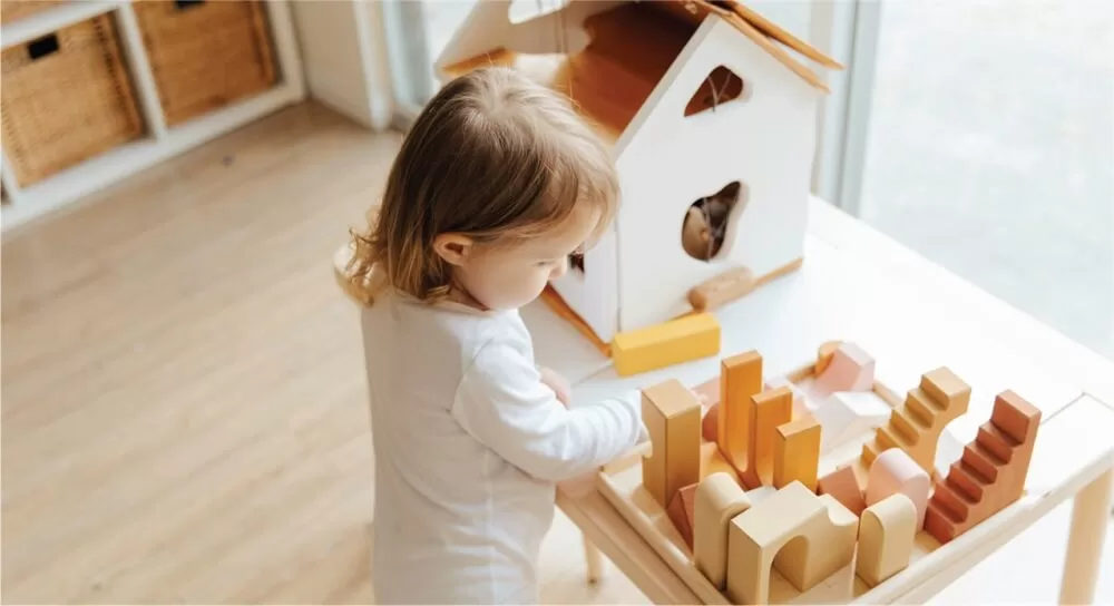 Niña pequeña jugando con bloques de construcción de madera en una mesa Montessori, mientras explora libremente en un ambiente preparado. A su lado, una casita de madera refuerza el juego simbólico y la autonomía.
