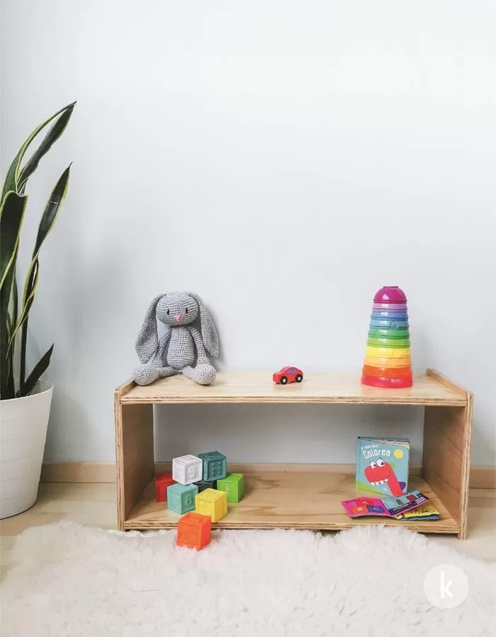 Shelf Montessori de madera con juguetes organizados a la altura del bebé: bloques sensoriales, libros y torre apilable, que fomentan la autonomía y el orden en el ambiente preparado.
