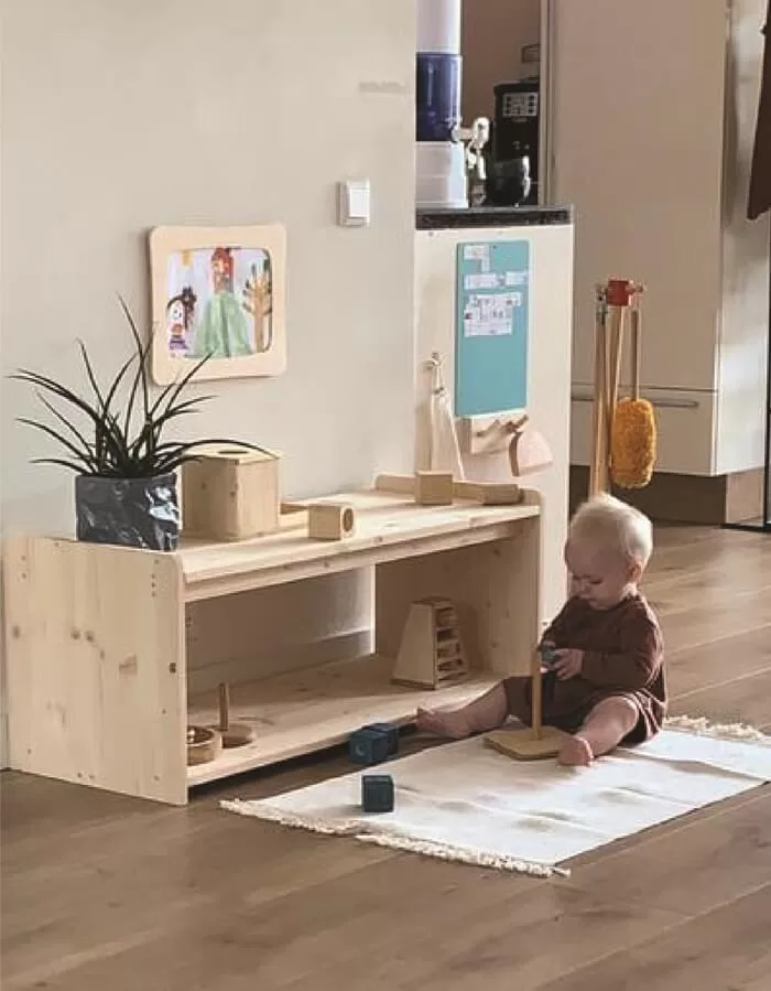 Bebe sobre alfombra juega con un juguete que ha tomado de estanteria montessori que se encuentra junto a el.