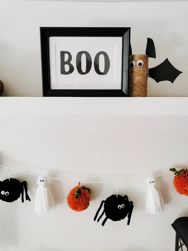 guirnalda-halloween-fantasmas-aranas-calabazas-artesanales Detalle de guirnalda artesanal de Halloween con fantasmas de hilo, calabazas de estambre y arañas negras bajo cuadro con la palabra BOO