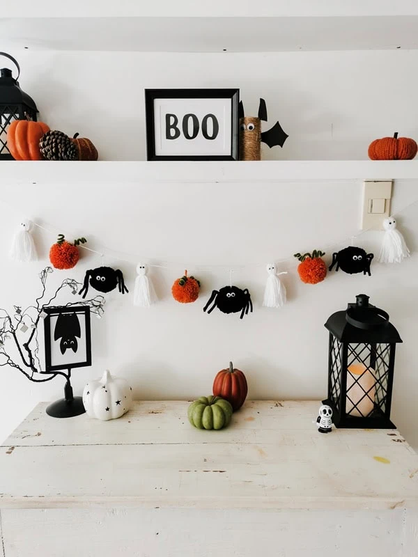guirnalda-halloween-fantasmas-calabazas-aranas-decoracion-mesa Guirnalda de Halloween con fantasmas, calabazas y arañas de estambre colgada debajo de una repisa con decoración otoñal.