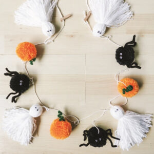 Guirnalda decorativa de Halloween hecha con pompones de hilo representando calabazas, arañas negras y fantasmas blancos, ideal para una decoración artesanal.