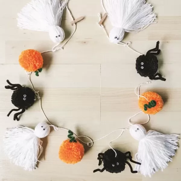 Guirnalda decorativa de Halloween hecha con pompones de hilo representando calabazas, arañas negras y fantasmas blancos, ideal para una decoración minimalista.
