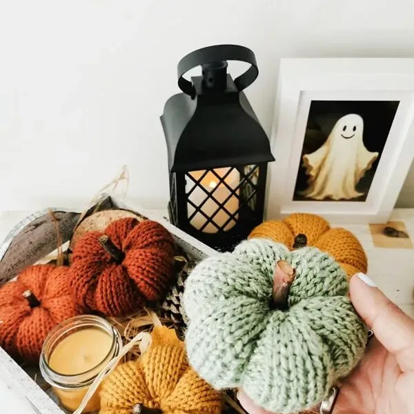 Calabaza tejida artesanal en tono verde sostenida en la mano, rodeada de otras calabazas decorativas de otoño.