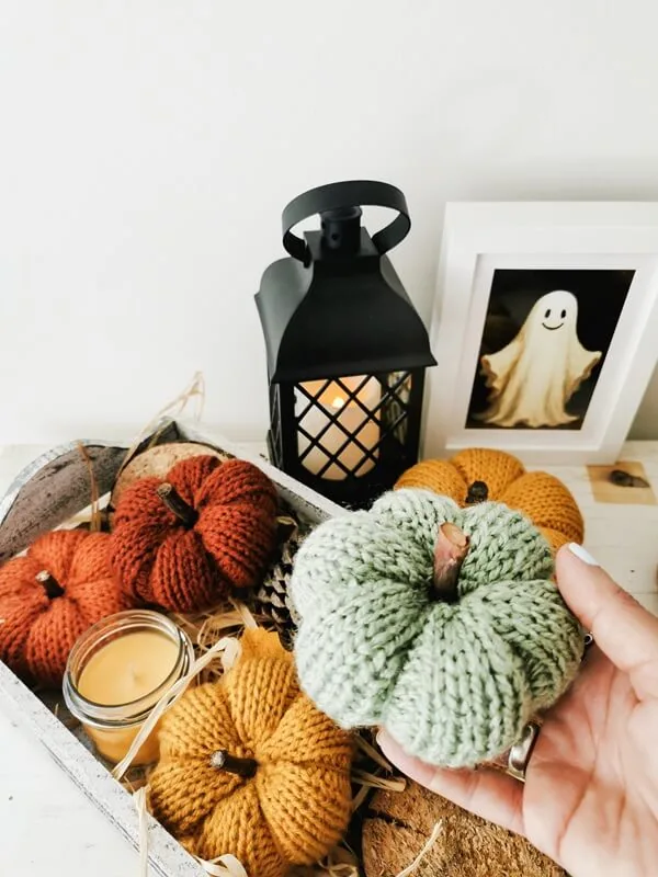 Calabaza tejida artesanal en tono verde sostenida en la mano, rodeada de otras calabazas decorativas de otoño.