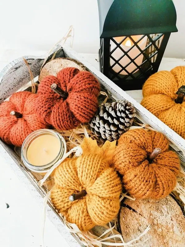 calabazas-tejidas-decoracion-otono-halloween Calabazas tejidas artesanales en tonos cálidos dentro de una bandeja decorativa con velas, piñas y hojas de otoño.